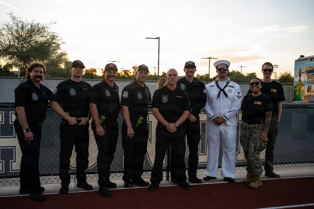 A group of approximately ten individuals pose for a photo at what appears to be a sports field or track during sunset. The group includes several firefighters in black uniforms with department patches, a U.S. Navy sailor in dress whites, and two individuals in Navy working uniforms. The setting appears to be an outdoor athletic facility, possibly during a community or military appreciation event.