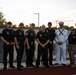 A group of approximately ten individuals pose for a photo at what appears to be a sports field or track during sunset. The group includes several firefighters in black uniforms with department patches, a U.S. Navy sailor in dress whites, and two individuals in Navy working uniforms. The setting appears to be an outdoor athletic facility, possibly during a community or military appreciation event.