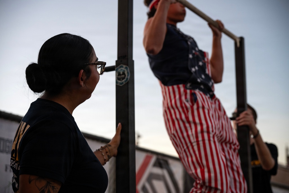 An outdoor fitness scene showing a person performing pull-ups on a metal bar while wearing American flag-themed clothing. A woman with glasses, a bun hairstyle, and visible tattoos observes from the left, possibly counting or spotting. A third person is partially visible on the right side of the frame.