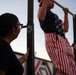 An outdoor fitness scene showing a person performing pull-ups on a metal bar while wearing American flag-themed clothing. A woman with glasses, a bun hairstyle, and visible tattoos observes from the left, possibly counting or spotting. A third person is partially visible on the right side of the frame.