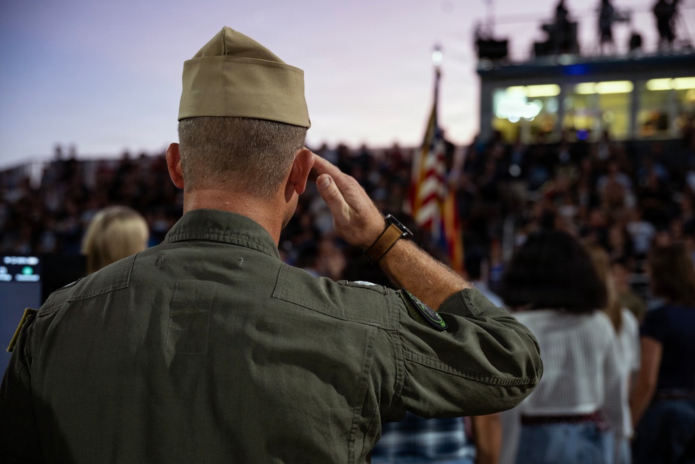A military service member in olive green flight suit and garrison cap renders a salute toward an American flag during what appears to be a public ceremony or sporting event. The scene is set at dusk in a stadium or outdoor venue filled with spectators. The image captures a moment of patriotic observance, likely during the National Anthem or a flag ceremony.