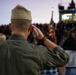 A military service member in olive green flight suit and garrison cap renders a salute toward an American flag during what appears to be a public ceremony or sporting event. The scene is set at dusk in a stadium or outdoor venue filled with spectators. The image captures a moment of patriotic observance, likely during the National Anthem or a flag ceremony.