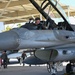 A pilot wearing a flight helmet marked with the number '69' is seated in the cockpit of an F-16 Fighting Falcon fighter jet with the canopy open. The aircraft is parked under a covered shelter (likely a flight line sunshade) at what appears to be a military air base. Ground crew personnel are visible in the background near other aircraft.