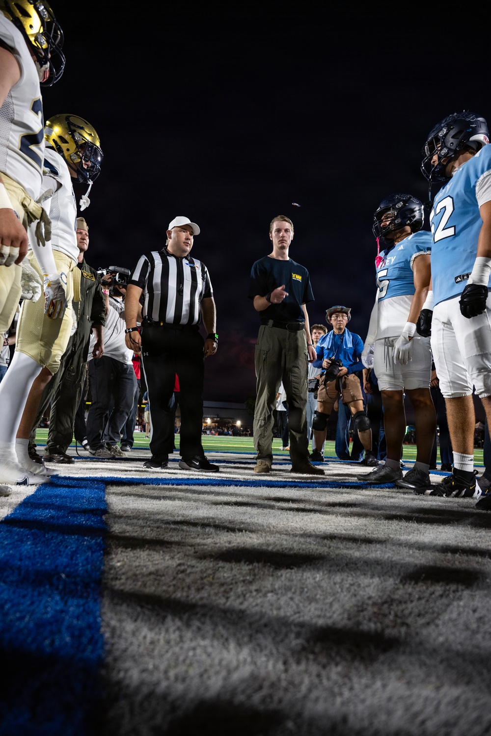 A nighttime American football game coin toss ceremony is taking place at midfield. A referee in a black-and-white striped shirt and white cap is officiating the toss, with players from two teams facing each other — one in white/gold uniforms and the other in light blue uniforms. A coin is visibly airborne in the upper portion of the frame, captured mid-flip.
