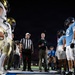 A nighttime American football game coin toss ceremony is taking place at midfield. A referee in a black-and-white striped shirt and white cap is officiating the toss, with players from two teams facing each other — one in white/gold uniforms and the other in light blue uniforms. A coin is visibly airborne in the upper portion of the frame, captured mid-flip.