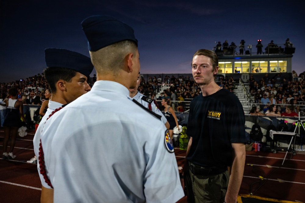 A nighttime outdoor event at a high school or college football stadium with a large crowd in the bleachers. Several individuals in white military-style uniforms with garrison caps, likely JROTC cadets, are visible on the track or field area. A young man in a dark Navy Special Operations t-shirt and olive pants appears to be interacting with or observing the uniformed cadets.