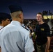A nighttime outdoor event at a high school or college football stadium with a large crowd in the bleachers. Several individuals in white military-style uniforms with garrison caps, likely JROTC cadets, are visible on the track or field area. A young man in a dark Navy Special Operations t-shirt and olive pants appears to be interacting with or observing the uniformed cadets.