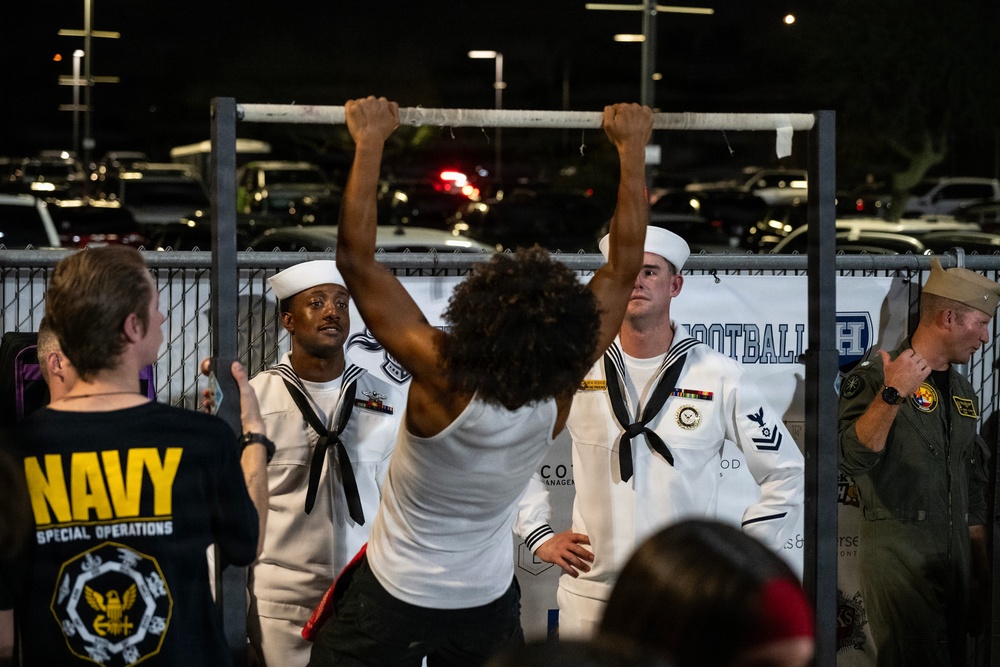 A nighttime outdoor event featuring U.S. Navy personnel in dress white uniforms observing a civilian performing pull-ups on a portable bar. The scene appears to be a Navy recruiting or public outreach event, possibly at a sporting venue given the parking lot setting and partial 'Football' signage visible in the background. Multiple Navy service members are present, including sailors in dress whites and at least one person in a flight suit.