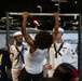 A nighttime outdoor event featuring U.S. Navy personnel in dress white uniforms observing a civilian performing pull-ups on a portable bar. The scene appears to be a Navy recruiting or public outreach event, possibly at a sporting venue given the parking lot setting and partial 'Football' signage visible in the background. Multiple Navy service members are present, including sailors in dress whites and at least one person in a flight suit.