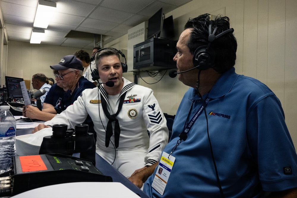 A U.S. Navy sailor in dress white uniform is being interviewed or conversing with a broadcaster wearing a blue AZPreps365 polo shirt, both wearing headsets. The setting appears to be a press box or broadcast booth at a state championship sporting event. Additional personnel are visible working in the background.