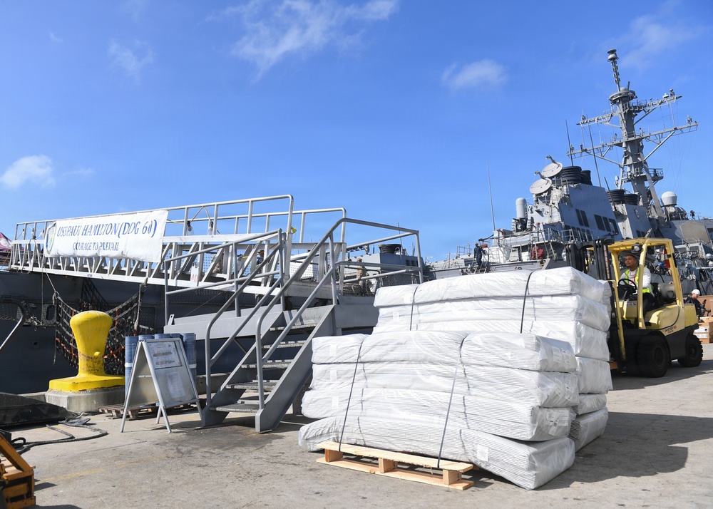 The image shows a U.S. Navy destroyer, USS Paul Hamilton (DDG-60), docked at a pier with cargo operations underway. A forklift operator is moving palletized bundles of white-wrapped materials on the dock. A banner on the gangway reads 'USS PAUL HAMILTON (DDG-60) COURAGE TO PREVAIL.'