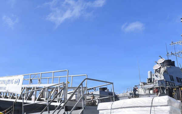 The image shows a U.S. Navy destroyer, USS Paul Hamilton (DDG-60), docked at a pier with cargo operations underway. A forklift operator is moving palletized bundles of white-wrapped materials on the dock. A banner on the gangway reads 'USS PAUL HAMILTON (DDG-60) COURAGE TO PREVAIL.'