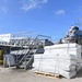 The image shows a U.S. Navy destroyer, USS Paul Hamilton (DDG-60), docked at a pier with cargo operations underway. A forklift operator is moving palletized bundles of white-wrapped materials on the dock. A banner on the gangway reads 'USS PAUL HAMILTON (DDG-60) COURAGE TO PREVAIL.'