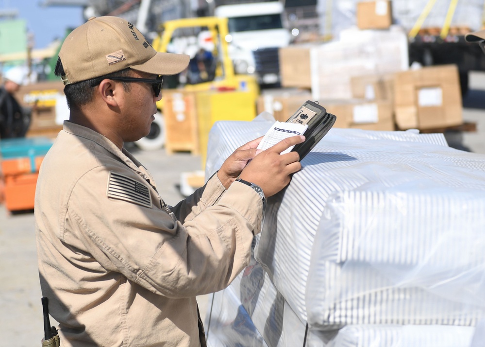 A U.S. military service member in tan/khaki uniform is scanning or checking documentation on a wrapped pallet at what appears to be a logistics or supply depot. The individual is using a handheld scanning device while referencing a paper document. The background shows a busy cargo area with forklifts, boxes, and additional palletized supplies.