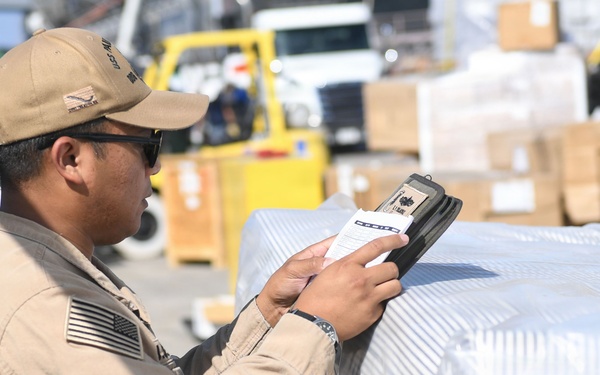 A U.S. military service member in tan/khaki uniform is scanning or checking documentation on a wrapped pallet at what appears to be a logistics or supply depot. The individual is using a handheld scanning device while referencing a paper document. The background shows a busy cargo area with forklifts, boxes, and additional palletized supplies.