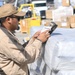 A U.S. military service member in tan/khaki uniform is scanning or checking documentation on a wrapped pallet at what appears to be a logistics or supply depot. The individual is using a handheld scanning device while referencing a paper document. The background shows a busy cargo area with forklifts, boxes, and additional palletized supplies.