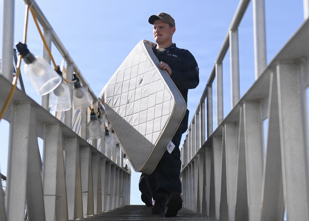 A U.S. Navy sailor in dark uniform and cap carries a mattress down an exterior metal stairway or gangway. The low-angle shot emphasizes the staircase structure and the sailor's upright posture while transporting the bedding. The setting appears to be a naval vessel or military facility.