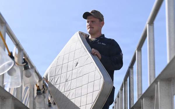 A U.S. Navy sailor in dark uniform and cap carries a mattress down an exterior metal stairway or gangway. The low-angle shot emphasizes the staircase structure and the sailor's upright posture while transporting the bedding. The setting appears to be a naval vessel or military facility.