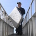 A U.S. Navy sailor in dark uniform and cap carries a mattress down an exterior metal stairway or gangway. The low-angle shot emphasizes the staircase structure and the sailor's upright posture while transporting the bedding. The setting appears to be a naval vessel or military facility.