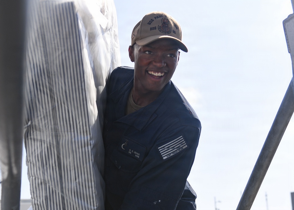 A smiling U.S. Navy sailor in a dark blue utility uniform is handling what appears to be a large wrapped or covered object. The sailor is wearing a tan USS ship cap and has a name tag and U.S. flag patch visible on the uniform. The image is taken from a low angle in an outdoor setting with bright sky in the background.