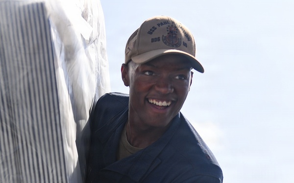 A smiling U.S. Navy sailor in a dark blue utility uniform is handling what appears to be a large wrapped or covered object. The sailor is wearing a tan USS ship cap and has a name tag and U.S. flag patch visible on the uniform. The image is taken from a low angle in an outdoor setting with bright sky in the background.