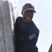 A smiling U.S. Navy sailor in a dark blue utility uniform is handling what appears to be a large wrapped or covered object. The sailor is wearing a tan USS ship cap and has a name tag and U.S. flag patch visible on the uniform. The image is taken from a low angle in an outdoor setting with bright sky in the background.