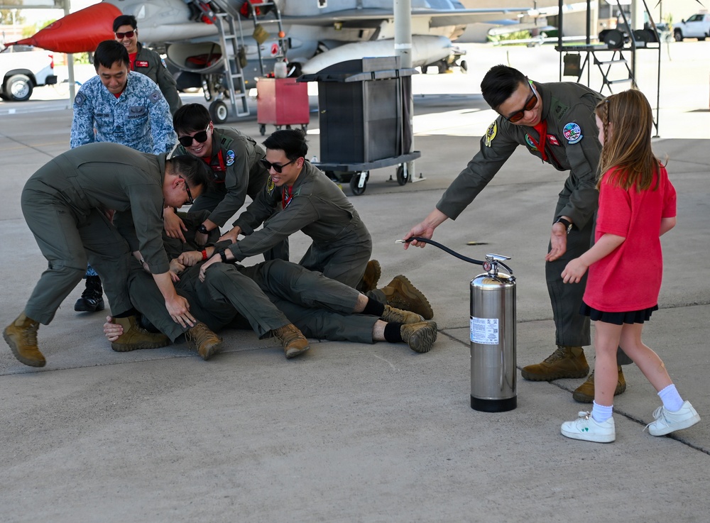 A group of military personnel in olive/sage green flight suits are engaged in what appears to be a lighthearted hazing or celebration ritual on an airfield tarmac, with one person being held down on the ground by colleagues. A young girl in a red shirt is standing nearby next to a stainless steel fire extinguisher, which one of the standing airmen is pointing toward the group. Military aircraft and ground equipment are visible in the background.