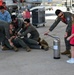 A group of military personnel in olive/sage green flight suits are engaged in what appears to be a lighthearted hazing or celebration ritual on an airfield tarmac, with one person being held down on the ground by colleagues. A young girl in a red shirt is standing nearby next to a stainless steel fire extinguisher, which one of the standing airmen is pointing toward the group. Military aircraft and ground equipment are visible in the background.
