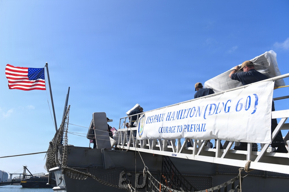 The image shows the USS Paul Hamilton (DDG 60), a U.S. Navy Arleigh Burke-class guided-missile destroyer, docked at a naval pier. A white banner displaying the ship's name, hull number, and motto 'Courage to Prevail' is prominently displayed on the gangway railing. Several personnel are visible on the ship's deck, with one appearing to carry a large wrapped item aboard.