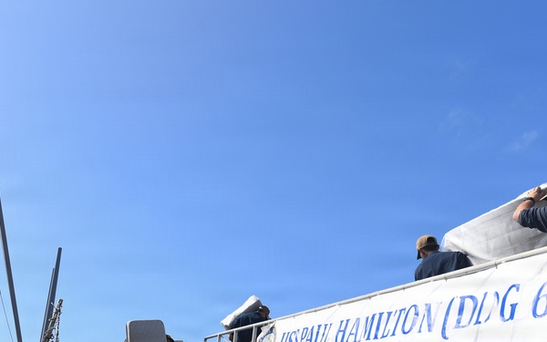 The image shows the USS Paul Hamilton (DDG 60), a U.S. Navy Arleigh Burke-class guided-missile destroyer, docked at a naval pier. A white banner displaying the ship's name, hull number, and motto 'Courage to Prevail' is prominently displayed on the gangway railing. Several personnel are visible on the ship's deck, with one appearing to carry a large wrapped item aboard.
