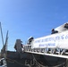 The image shows the USS Paul Hamilton (DDG 60), a U.S. Navy Arleigh Burke-class guided-missile destroyer, docked at a naval pier. A white banner displaying the ship's name, hull number, and motto 'Courage to Prevail' is prominently displayed on the gangway railing. Several personnel are visible on the ship's deck, with one appearing to carry a large wrapped item aboard.