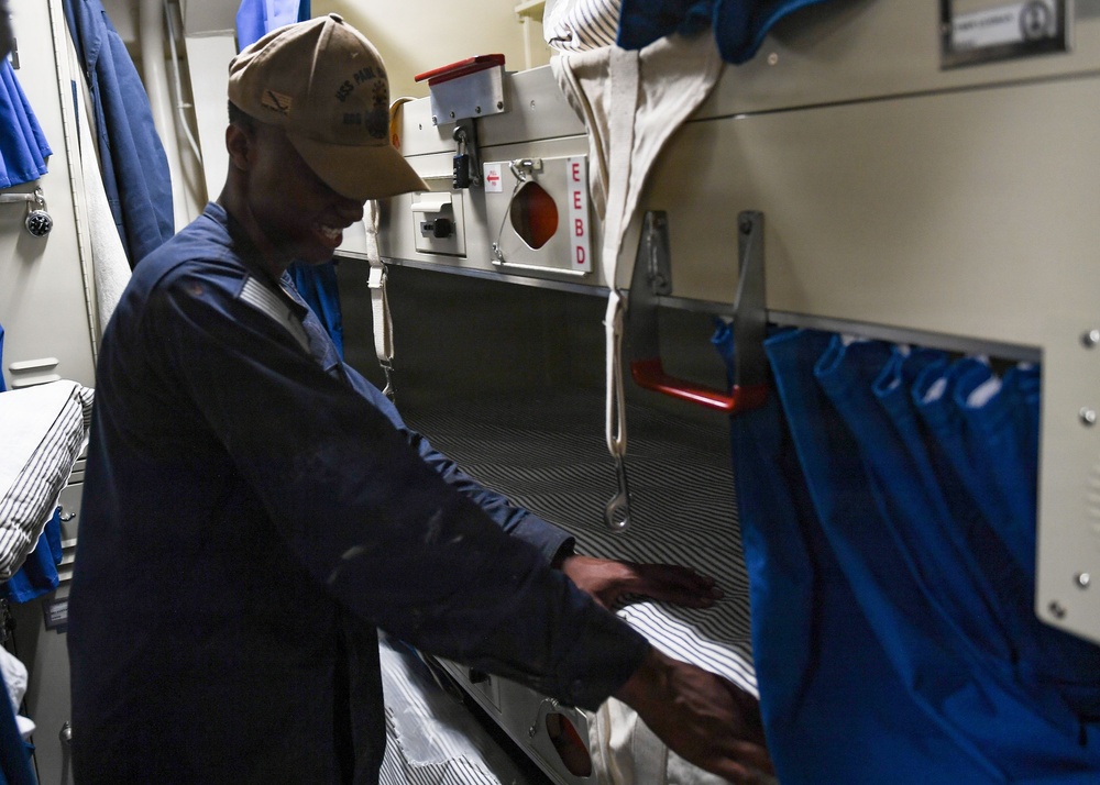 A person wearing a navy blue coverall uniform and a tan baseball cap is standing in a tight berthing compartment aboard a military naval vessel. They appear to be smiling and gesturing toward a rack (bunk bed) with blue privacy curtains. The space shows typical U.S. Navy shipboard berthing features including stacked racks, an EEBD (Emergency Escape Breathing Device) mounted on the wall, and limited space.