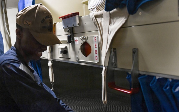 A person wearing a navy blue coverall uniform and a tan baseball cap is standing in a tight berthing compartment aboard a military naval vessel. They appear to be smiling and gesturing toward a rack (bunk bed) with blue privacy curtains. The space shows typical U.S. Navy shipboard berthing features including stacked racks, an EEBD (Emergency Escape Breathing Device) mounted on the wall, and limited space.