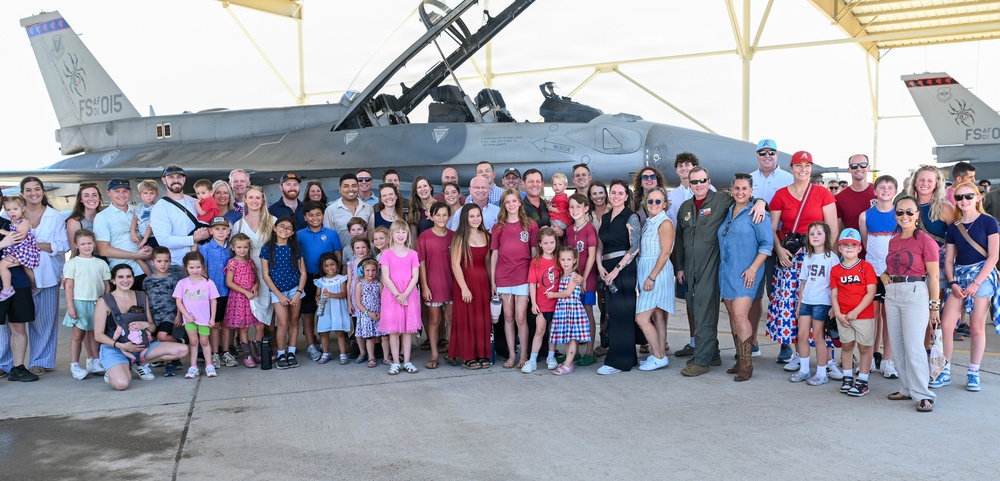 A large group of military personnel, families, and children pose for a group photo in front of an F-16 Fighting Falcon jet aircraft inside a hangar or covered tarmac area. The gathering appears to be a military family day or homecoming event, with participants dressed in casual and patriotic attire. At least one individual is wearing a flight suit, suggesting an active-duty military context.