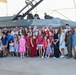 A large group of military personnel, families, and children pose for a group photo in front of an F-16 Fighting Falcon jet aircraft inside a hangar or covered tarmac area. The gathering appears to be a military family day or homecoming event, with participants dressed in casual and patriotic attire. At least one individual is wearing a flight suit, suggesting an active-duty military context.