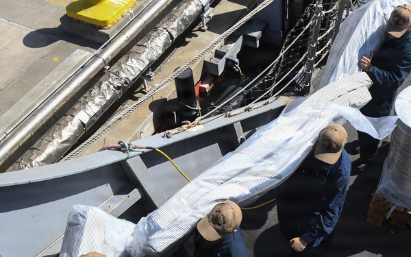 An aerial/overhead view of several personnel in dark blue uniforms and tan/khaki caps working on what appears to be a naval vessel deck. They are handling large wrapped or covered items, possibly equipment or parts being loaded or unloaded. A pallet with stacked materials is visible to the right side of the image.