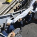 An aerial/overhead view of several personnel in dark blue uniforms and tan/khaki caps working on what appears to be a naval vessel deck. They are handling large wrapped or covered items, possibly equipment or parts being loaded or unloaded. A pallet with stacked materials is visible to the right side of the image.