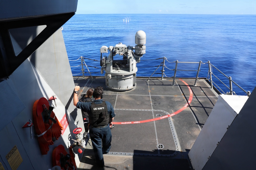 Two US Navy personnel are standing on the deck of a naval vessel observing a weapon system, likely a Phalanx Close-In Weapon System (CIWS), which appears to be firing or has recently fired, evidenced by smoke visible near the barrel. The deck is marked with red 'DANGER AREA' warnings painted in a circular pattern around the weapon mount. The vessel is operating in open blue ocean waters under clear skies.