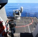Two US Navy personnel are standing on the deck of a naval vessel observing a weapon system, likely a Phalanx Close-In Weapon System (CIWS), which appears to be firing or has recently fired, evidenced by smoke visible near the barrel. The deck is marked with red 'DANGER AREA' warnings painted in a circular pattern around the weapon mount. The vessel is operating in open blue ocean waters under clear skies.