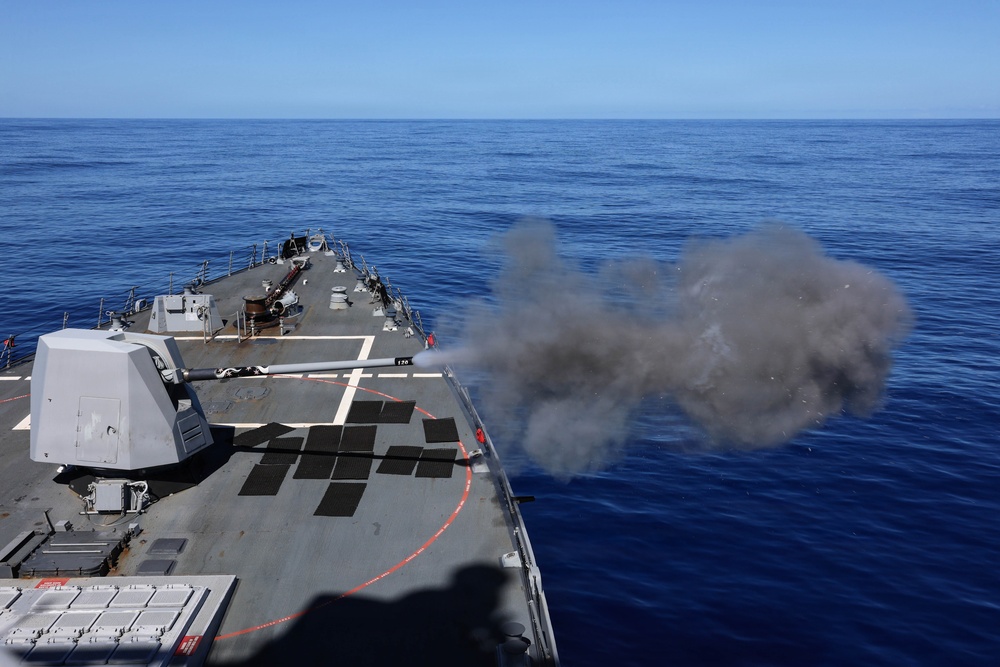 A naval warship is firing its forward deck gun during what appears to be a live-fire exercise at sea. A large cloud of gray smoke and gun blast debris is visible at the muzzle of the cannon, which is pointed to the starboard side. The vessel is underway on calm, deep blue open ocean under clear skies.