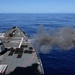 A naval warship is firing its forward deck gun during what appears to be a live-fire exercise at sea. A large cloud of gray smoke and gun blast debris is visible at the muzzle of the cannon, which is pointed to the starboard side. The vessel is underway on calm, deep blue open ocean under clear skies.