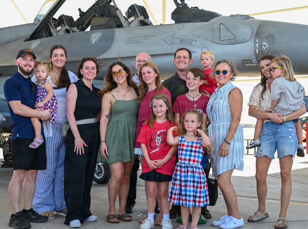 A large group of adults and children pose together in front of a military jet aircraft, likely an F-16 Fighting Falcon, on what appears to be a flight line or tarmac. The gathering appears to be a family or group photo opportunity at a military air base, possibly during an airshow or homecoming event. The mood is celebratory and casual, with people smiling and dressed in everyday civilian clothing.