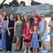 A large group of adults and children pose together in front of a military jet aircraft, likely an F-16 Fighting Falcon, on what appears to be a flight line or tarmac. The gathering appears to be a family or group photo opportunity at a military air base, possibly during an airshow or homecoming event. The mood is celebratory and casual, with people smiling and dressed in everyday civilian clothing.
