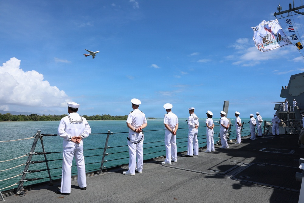 Naval personnel in white dress uniforms are standing at attention along the deck railing of a warship, appearing to man the rails in a formal ceremonial formation. A commercial aircraft is visible flying overhead, and the ship appears to be in a tropical coastal setting with turquoise waters and lush green vegetation visible in the background. Signal flags and what appears to be a naval ensign are flying from the ship's mast in the upper right.