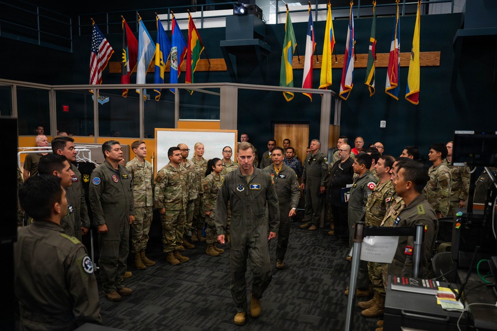 A military briefing or gathering is taking place in what appears to be a command center or operations room, with a senior officer in a flight suit walking through a formation of multinational military personnel. Multiple national flags are displayed prominently in the background, suggesting an international or joint military exercise. Personnel are wearing various uniforms including US Air Force flight suits and Army combat uniforms, as well as uniforms from other nations.