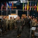 A military briefing or gathering is taking place in what appears to be a command center or operations room, with a senior officer in a flight suit walking through a formation of multinational military personnel. Multiple national flags are displayed prominently in the background, suggesting an international or joint military exercise. Personnel are wearing various uniforms including US Air Force flight suits and Army combat uniforms, as well as uniforms from other nations.