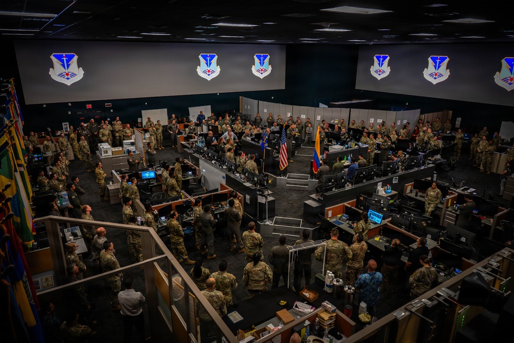 A large military operations center or command facility filled with hundreds of personnel in camouflage uniforms gathered for what appears to be a formal briefing or ceremony. The room features multiple workstations with computer monitors, large display screens showing a military unit insignia, and national flags including the United States flag and what appears to be a Colombian flag. The elevated vantage point provides a wide overhead view of the entire floor.