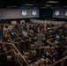 A large military operations center or command facility filled with hundreds of personnel in camouflage uniforms gathered for what appears to be a formal briefing or ceremony. The room features multiple workstations with computer monitors, large display screens showing a military unit insignia, and national flags including the United States flag and what appears to be a Colombian flag. The elevated vantage point provides a wide overhead view of the entire floor.