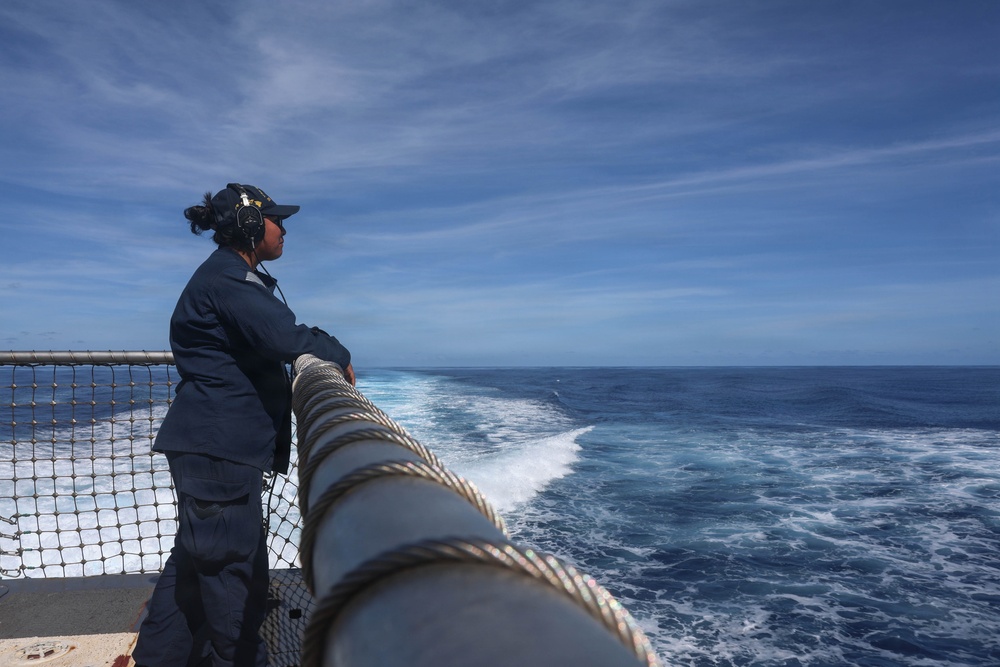 A naval or coast guard service member in dark blue coveralls stands at the stern railing of a military vessel, looking out over the open ocean. The ship's wake is clearly visible trailing behind, indicating the vessel is underway at sea. The scene is captured under a partly cloudy blue sky with calm to moderate sea conditions.