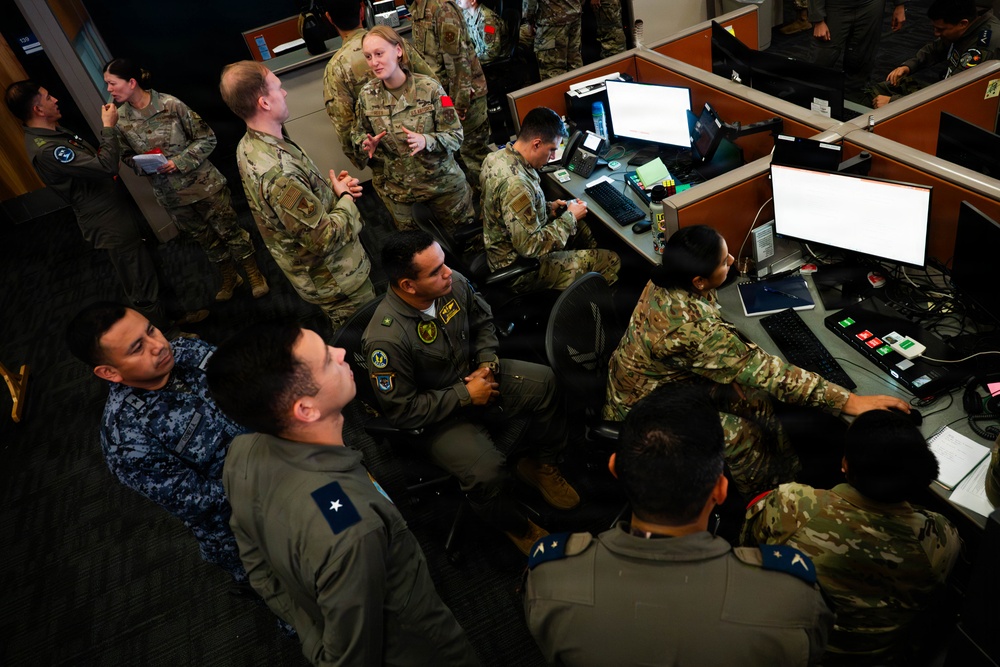 A group of military personnel from multiple branches and possibly allied nations are gathered in what appears to be a command operations center or joint operations facility. Personnel are wearing various uniforms including US Army/Air Force OCP camouflage, Navy digital camouflage, and olive/sage flight suits with rank insignia and unit patches. The scene suggests a briefing, training exercise, or joint operational planning session at workstations equipped with multiple computer monitors.